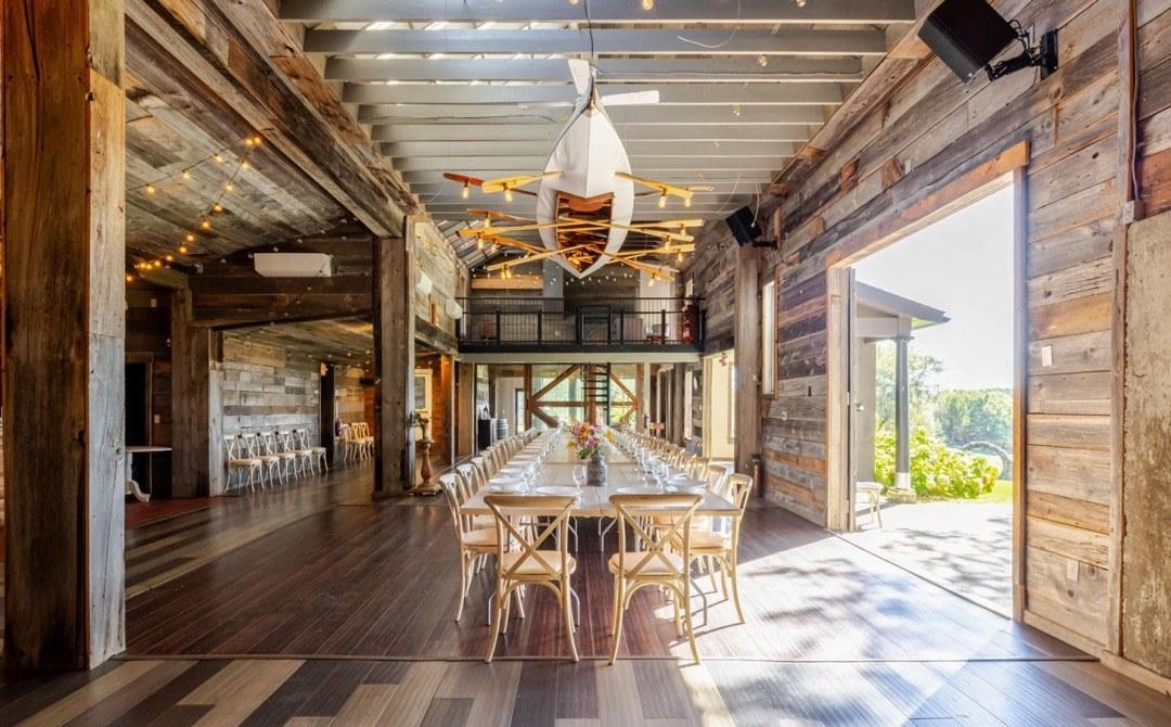Interior of the restored barn at Twelve Ponds with weathered wood walls, a long farmhouse table set for dinner, and open doors leading to the lawn.