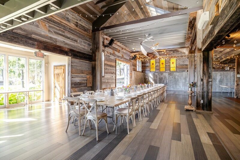 A long harvest table set in the barn at Twelve Ponds, cross-back chairs, string lights and vaulted reclaimed ceiling.
