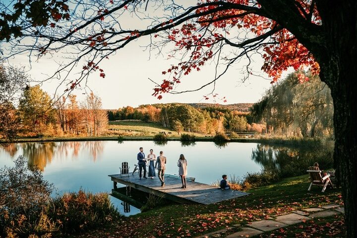 A ceremony on the wooden dock at Twelve Ponds at sunset, red maple leaves overhead.
