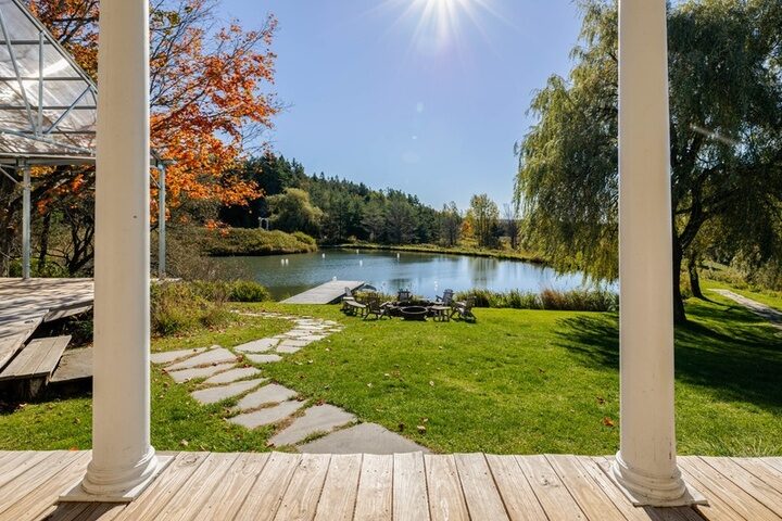 View through the farmhouse porch columns toward the pond, stone path, fire pit and Adirondack chairs.