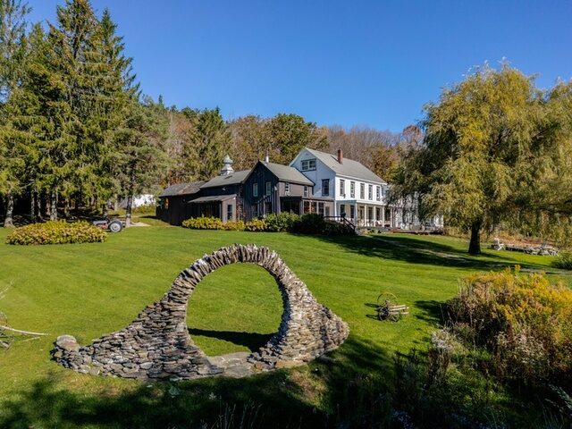 The stone moon-gate arch on the meadow at Twelve Ponds, framed between the farmhouse and dark barn.